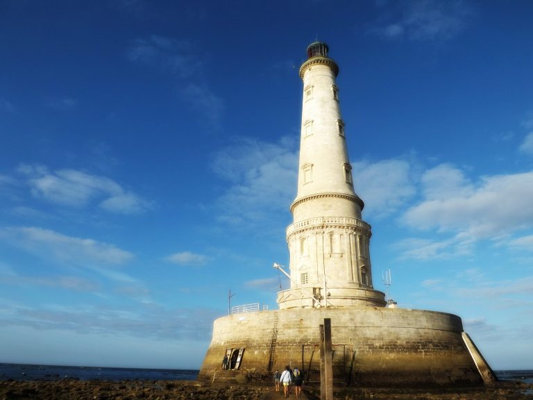A white stone lighthouse nestled on a round three storey round base rises into a deep blue september sky.Tourists are mingling around the lighthouse and base which is accesible because of low tide.