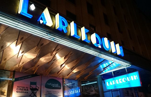 Photo nocturne de l'entrée du cinéma L'Arlequin. Son nom s'affiche en néon bleu au-dessus de la porte. Des affiches pour le festival du cinéma nigérian de 2023, la semaine Nollywood et des films français sont visibles dans la vitre en dessous. Le bâtiment semble doré.   