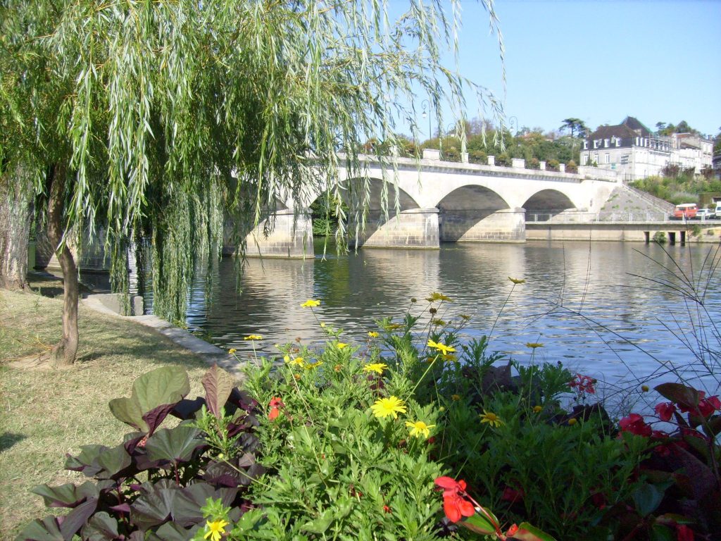 A stone bridge with multiple arches spans a calm river. In the foreground, colourful flowers and green plants grow next to the water. A willow tree’s branches hang over the riverbank. Houses and trees are visible on a sunny day in the background.