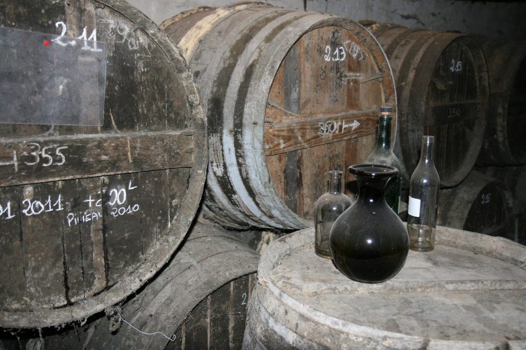 Old wooden barrels, some marked with white chalk writing, are stacked in a dim cellar. On one barrel in the foreground, there are four glass bottles—one dark, three clear—of various shapes and sizes. The scene suggests wine or spirit ageing.